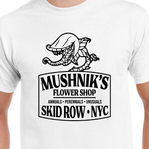 Puede incluir: Camiseta blanca con una ilustraci&oacute;n en blanco y negro de una planta carn&iacute;vora y el texto "Mushnik's Flower Shop. Annuals - Perennials - Unusuasls. Skid Row. NYC."