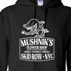 Puede incluir: Sudadera con capucha negra con un gr&aacute;fico blanco de una planta carn&iacute;vora y el texto "Mushnik's Flower Shop, Annuals - Perennials - Unusuasls, Skid Row - NYC."
