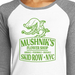 Puede incluir: Una camiseta de b&eacute;isbol blanca y gris con un gr&aacute;fico verde de una planta carn&iacute;vora y el texto "Mushnik's Flower Shop, Annuals - Perennials - Unusuasls, Skid Row - NYC."