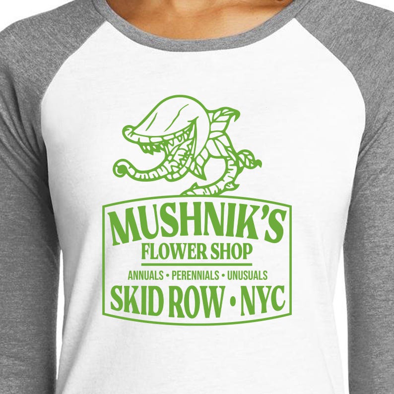 Puede incluir: Una camiseta de b&eacute;isbol blanca y gris con un gr&aacute;fico verde de una planta carn&iacute;vora y el texto "Mushnik's Flower Shop, Annuals - Perennials - Unusuasls, Skid Row - NYC."