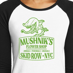 Puede incluir: Una camiseta de b&eacute;isbol blanca y negra con una ilustraci&oacute;n verde de una planta carn&iacute;vora y el texto "Mushnik's Flower Shop. Annuals - Perennials - Unusuasls. Skid Row. NYC."