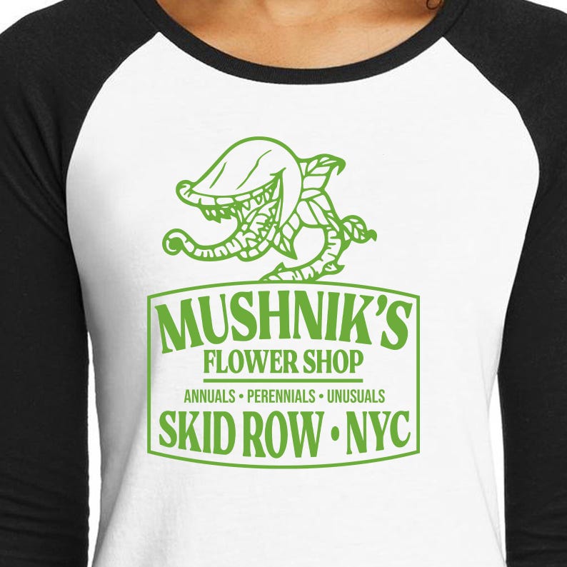 Puede incluir: Una camiseta de b&eacute;isbol blanca y negra con una ilustraci&oacute;n verde de una planta carn&iacute;vora y el texto "Mushnik's Flower Shop. Annuals - Perennials - Unusuasls. Skid Row. NYC."