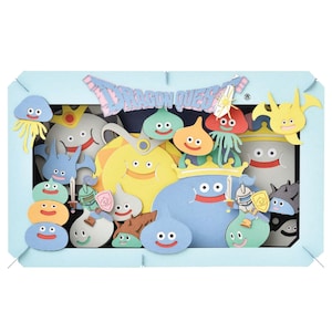 以下が含まれることがあります： 明るい青色のフレームの中に、笑顔のスライムやその他の生き物など、さまざまなカラフルな漫画のキャラクターが登場するペーパーシアター。「DRAGON QUEST」と書かれたバナー。www.CraftWithLava.comをご覧ください。