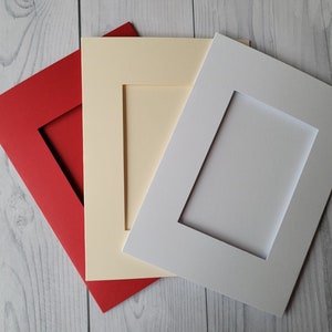 May include: Three rectangular picture mounts in red, cream, and white. Each mount has a rectangular aperture in the centre. The mounts are arranged diagonally, overlapping, against a light grey wood grain background.
