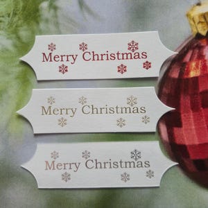May include: Three white rectangular Christmas gift tags with the words "Merry Christmas" printed in red, gold, and bronze, along with snowflake designs. A red and gold Christmas bauble is in the background.