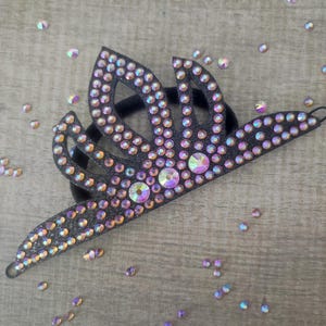 May include: A black tiara-style headband adorned with iridescent rhinestones. The tiara features a central design with three arched sections, all covered in sparkling gems. Scattered iridescent rhinestones surround the headband on a wooden surface.