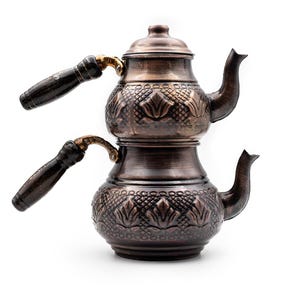 Peut inclure: Une théière à deux niveaux de couleur cuivrée, ornée de motifs floraux et d'un couvercle. Elle possède deux becs verseurs et deux poignées en bois avec des accents dorés. La théière est probablement utilisée pour infuser du thé.