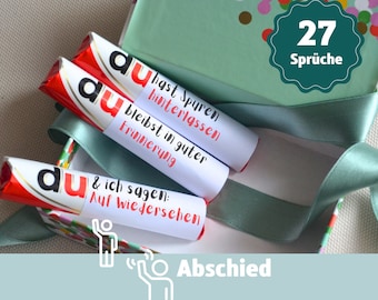 May include: A box with a green and white confetti pattern lid, filled with chocolate bars wrapped in red and white paper. The chocolate bars have white labels with farewell messages in German. The box is tied with a green ribbon. The lid has a green circle with the text "27 Sprüche" in white.