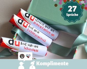 May include: A green box with confetti print and a green ribbon. The box contains chocolate bars wrapped in red foil with white labels that say "du" and have a variety of compliments written on them. The box lid has a green circle with the text "27 Sprüche" on it. The text "Komplimente" is written on a light blue background below the box.