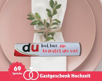 May include: A white napkin with a green sprig tied with twine, a chocolate bar with the text "du" in red and black, and a white label with the text "bist hier, das bedeutet uns viel". The chocolate bar is on a pink plate. The image also includes a red circle with the text "69 Sprüche" and a red ring with a white diamond.