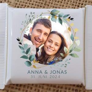 Puede incluir: Barra de chocolate blanco con una foto de una pareja en una corona floral verde y dorada. El texto "ANNA & JONAS 31. JUNI 2024" está impreso debajo de la foto.