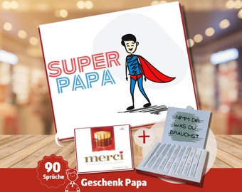 May include: A red and white card with the text "Super Papa" and a cartoon image of a superhero. The card is next to a box of Merci chocolates and a box of chocolate bars with the text "Nimm dir was du brauchst".