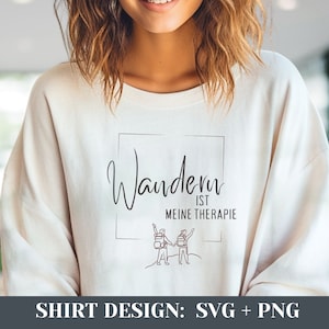 以下が含まれることがあります： バックパックを背負ったハイキングをしている2人の人物が描かれたグラフィックデザインの白いTシャツ。テキスト「Wandern ist meine Therapie」は筆記体で書かれています。