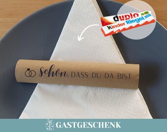 May include: A brown paper tube with the text "Schön, dass du da bist" and a small illustration of two rings. The tube is sitting on a white napkin with a small chocolate bar in the background.