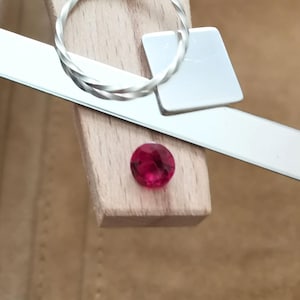 May include: A faceted, round, deep red gemstone sits on a wooden surface, next to a silver metal ring and a rectangular metal piece. The ring has a twisted design. A long, flat metal tool is also present, with a light brown background.