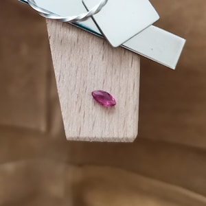 May include: A marquise-cut, deep pink gemstone rests on a wooden jeweler's bench. A silver ring and metal tools are in the background. The gemstone is the focal point, showcasing its vibrant color and shape, suggesting jewelry making or design.