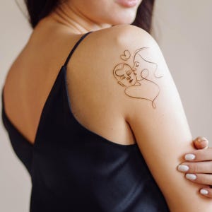 May include: A line art tattoo design of a mother and child embracing, with a heart above their heads. The tattoo is on a person's shoulder.