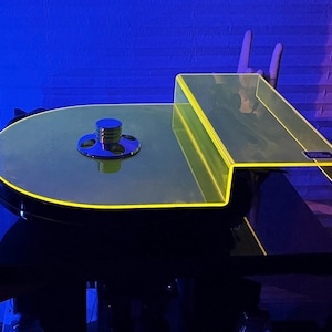 Op de afbeelding: Een moderne platenspeler met een neongele acryl accent. De platenspeler heeft een cirkelvormige schotel met een chromen midden en een bijpassende neongele plank. De basis is zwart en het ontwerp is strak en eigentijds.