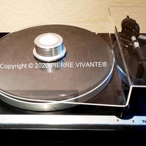 Puede incluir: Un tocadiscos Thorens TD 320 negro con una cubierta antipolvo de acrílico transparente. El tocadiscos tiene un plato plateado y un brazo de lectura negro. El tocadiscos está sobre una superficie negra. Copyright © 2020 PIERRE VIVANTE®