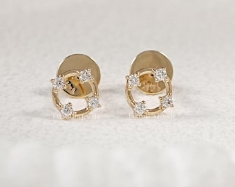 May include: A pair of gold-colored stud earrings. Each earring features a square design with four clear, round gemstones set within the square. The earrings are displayed on a white surface.