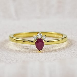 May include: A gold ring featuring a pear-shaped red ruby gemstone at the centre, accented by three small clear round stones arranged in a crown-like setting above the ruby. The text EverGlam Jewels is visible on the right side of the ring band.