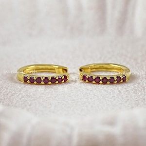 May include: A pair of gold hoop earrings featuring a row of small, round, deep red rubies set along the front curve. The text EverGlam Jewels is visible on each earring. These circular earrings have a polished finish and a hinged closure mechanism.