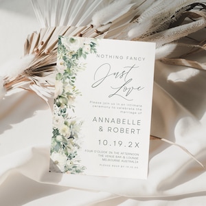 May include: A white wedding invitation with green foliage and white flowers on the left side. The text reads "Nothing Fancy Just Love" with the couple's names and wedding date. The invitation is on a white background with a dried palm leaf.