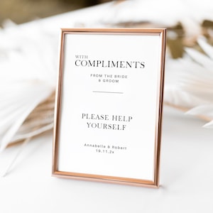 May include: A framed sign with a rose gold border. The sign reads "WITH COMPLIMENTS FROM THE BRIDE & GROOM PLEASE HELP YOURSELF" with the names Annabelle & Robert and the date 19.11.2x. The background is blurred with white decorative elements.