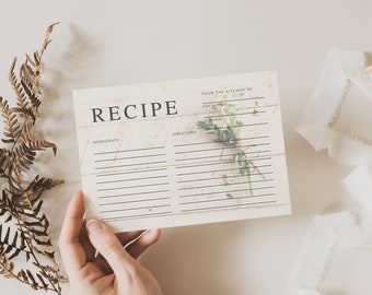 May include: A vintage-style recipe card with the word "RECIPE" at the top. The card has lines for ingredients and directions, with a sprig of greenery. A hand holds the card, with a white ribbon and dried fern in the background.