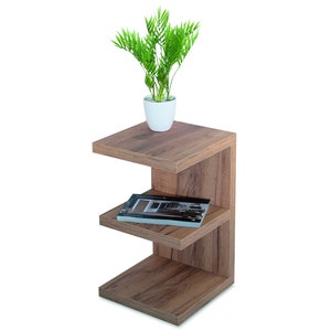 Puede incluir: Una mesa auxiliar de madera con tres estantes. El estante superior tiene una peque&ntilde;a planta en maceta con hojas verdes. El estante del medio tiene un libro encima. La mesa es marr&oacute;n y tiene un dise&ntilde;o moderno.