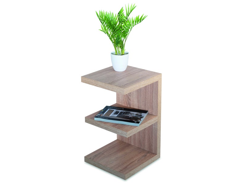 Puede incluir: Una mesa auxiliar de madera marr&oacute;n claro con tres estantes. El estante superior tiene una peque&ntilde;a maceta blanca con una planta verde. Un libro est&aacute; en el estante del medio.