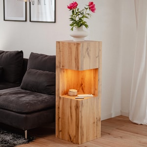 May include: A tall, wooden display cabinet with a built-in shelf illuminated by warm light. A white vase with pink flowers sits on top. The cabinet has a natural wood grain finish and a modern design.