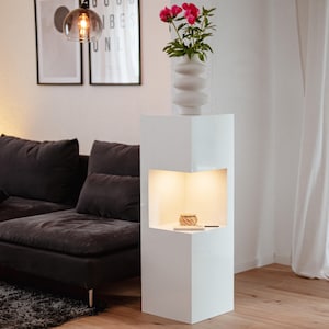 Op de afbeelding: Een moderne witte displaystandaard met een ingebouwde verlichte nis. Bovenop staat een witte vaas met roze bloemen. De standaard staat in een woonkamer met een donkergrijze bank en kunstwerken op de achtergrond.