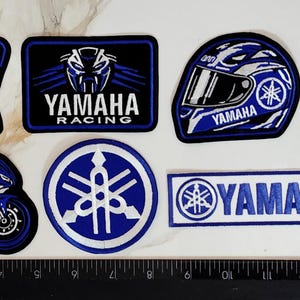 May include: A collection of eight embroidered Yamaha Racing patches in blue, black, and white, featuring logos, a motorcycle, a helmet, and the number 46, arranged next to a ruler showing a length of approximately 16 inches.