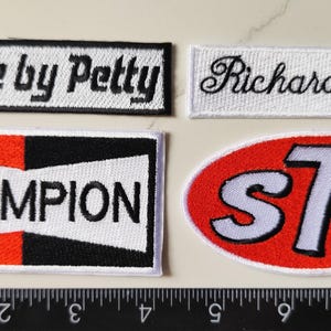 Puede incluir: Cuatro parches bordados con texto. Los parches incluyen "Dodge by Petty", "Richard Petty", "CHAMPION" y un parche ovalado con "STP". Los parches son negros, blancos, rojos y naranjas.