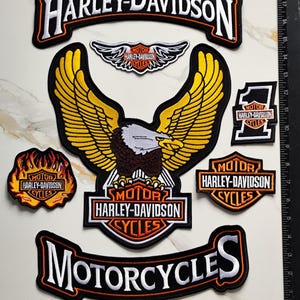 Op de afbeelding: Een verzameling geborduurde Harley-Davidson patches, waaronder een groot adelaarembleem, een gebogen bovenste rocker, een onderste rocker met de tekst MOTORCYCLES, een vlamvormige patch, een nummer één patch en een klein gevleugeld logo naast een liniaal van 48 cm.