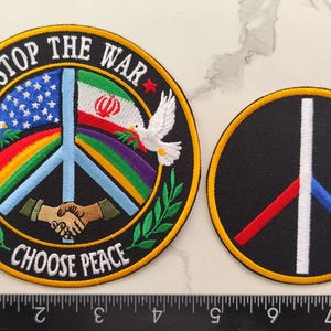 Könnte beinhalten: Zwei gestickte Aufnäher. Einer ist rund mit schwarzem Hintergrund und gelbem Rand. Er zeigt ein Friedenssymbol, einen Regenbogen, die amerikanische und iranische Flagge, Tauben und die Texte "STOP THE WAR" und "CHOOSE PEACE". Der andere Aufnäher ist ein Friedenssymbol mit roten, weißen und blauen Streifen.