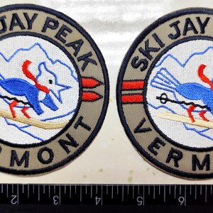 May include: Two circular embroidered patches. Each patch features a blue bird skiing with a red scarf, the text "SKI JAY PEAK VERMONT" in a circular design, and two red arrows. The patches are tan, blue, red, and white.