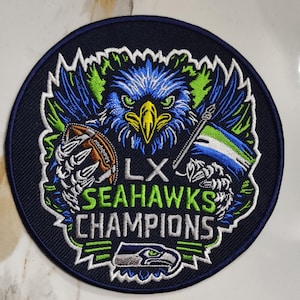 May include: A circular patch featuring a detailed eagle design in blue, green, and yellow, with a football and flag. The text reads "LX SEAHAWKS CHAMPIONS" in white and green. A smaller Seahawks logo is at the bottom.