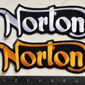 Peut inclure: Deux écussons brodés affichant le mot Norton dans une police stylisée. Un écusson présente des lettres blanches sur fond noir, et l'autre des lettres jaunes sur fond noir. Une règle indique que les écussons mesurent environ 15 cm de large.