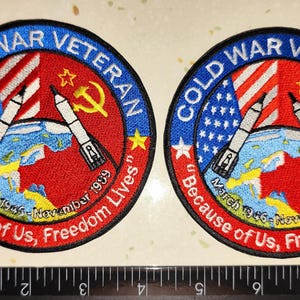 Puede incluir: Dos parches bordados circulares con borde azul y la inscripción "COLD WAR VETERAN". Los parches muestran la bandera estadounidense, misiles, una hoz y un martillo, y el texto "Because of Us, Freedom Lives."