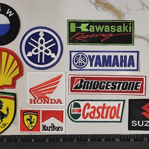 Op de afbeelding: Een verzameling van twaalf geborduurde patches met diverse logo's van auto- en motormerken, waaronder BMW, Yamaha, Kawasaki Racing, Shell, Honda, Bridgestone, Castrol, Ferrari, Marlboro en Suzuki, gerangschikt naast een liniaal van 35 cm.