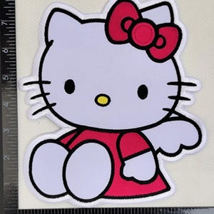 May include: Embroidered patch of Hello Kitty. The patch shows Hello Kitty in a seated pose, wearing a red dress and a matching bow. The design includes black outlines and a yellow nose, set against a white background.