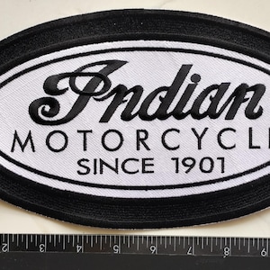 Op de afbeelding: Een ovaalvormige geborduurde patch met een zwarte rand en een wit midden. De tekst Indian MOTORCYCLE SINCE 1901 is in zwart borduurwerk aangebracht. Een liniaal langs de linker- en onderkant toont dat de patch ongeveer 26,7 cm breed is.