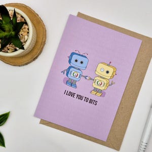 Puede incluir: Tarjeta de felicitación lila con dos robots de dibujos animados tomados de la mano. El texto dice "I LOVE YOU TO BITS". Los robots son azules y amarillos. La tarjeta está sobre un sobre marrón.
