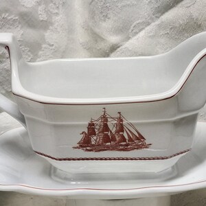 May include: White ceramic gravy boat with a matching saucer. The gravy boat features a handle and a spout, with a brown illustration of a sailing ship on the side. The rim of the boat and saucer are trimmed in brown.