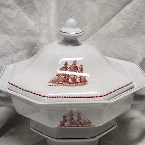 May include: White ceramic serving dish with a lid, featuring a red illustration of a sailing ship. The dish has a geometric shape with a handle on each side. The rim and lid are trimmed with a red dotted pattern.