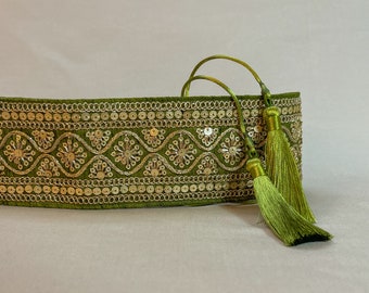 Può includere: Una cintura in velluto verde con paillettes dorate e nappe. La cintura è decorata con un motivo ripetuto di piccoli cerchi e fiori.