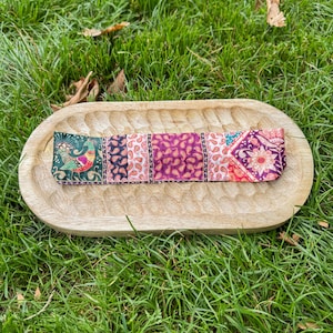 Può includere: Una fascia colorata con un design patchwork con motivi paisley e floreali. La fascia è esposta su un vassoio di legno marrone chiaro, su uno sfondo di erba verde.