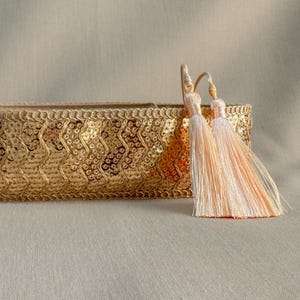 Pode incluir: Uma caixa decorativa retangular dourada com uma superfície texturizada com lantejoulas. Duas borlas cor de pêssego pendem do canto superior direito. A caixa tem uma guarnição decorativa ao longo da borda superior.
